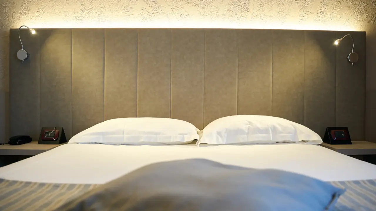 Una camera del First Hotel la sera: la testata imbottita illuminata dall’alto, due lampade da lettura accese e i cuscini bianchi