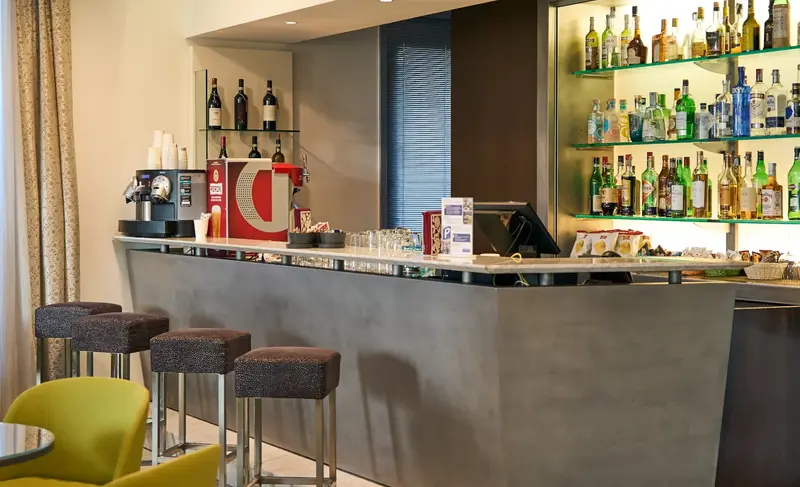 Il lobby bar: il bancone grigio con gli sgabelli e la parete delle bottiglie illuminata di verde
