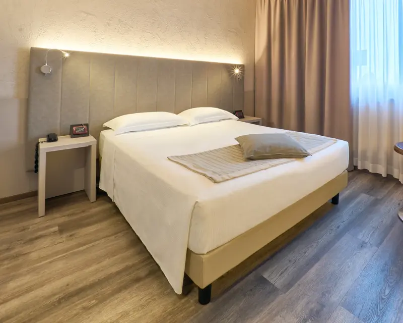 La camera Deluxe: letto matrimoniale con la testata imbottita illuminata dall'alto, coperta a righe e finestra con le tende