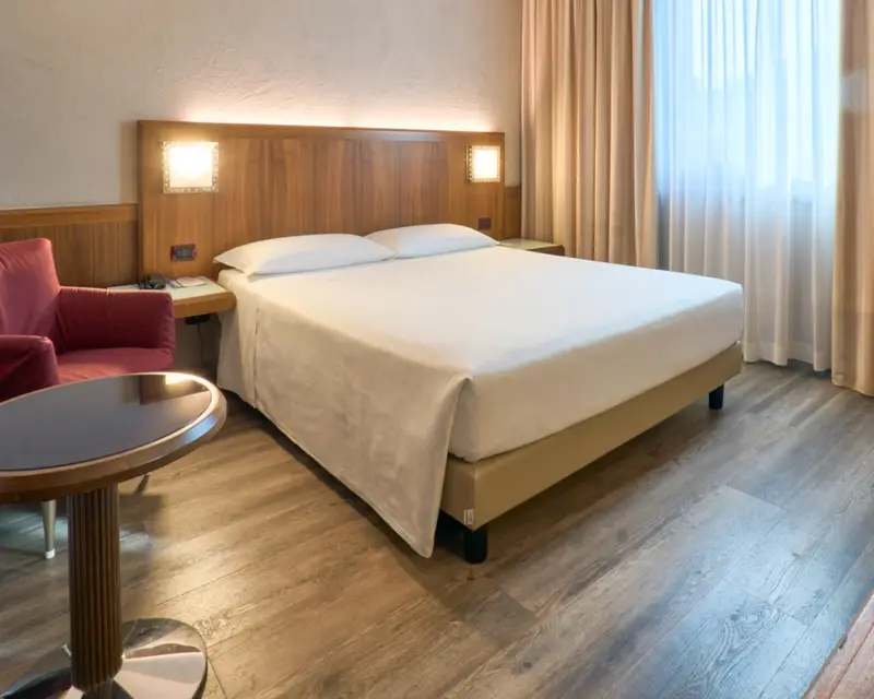 La camera Standard: letto matrimoniale, testata in legno con le luci accese, poltrona rossa e tende chiare
