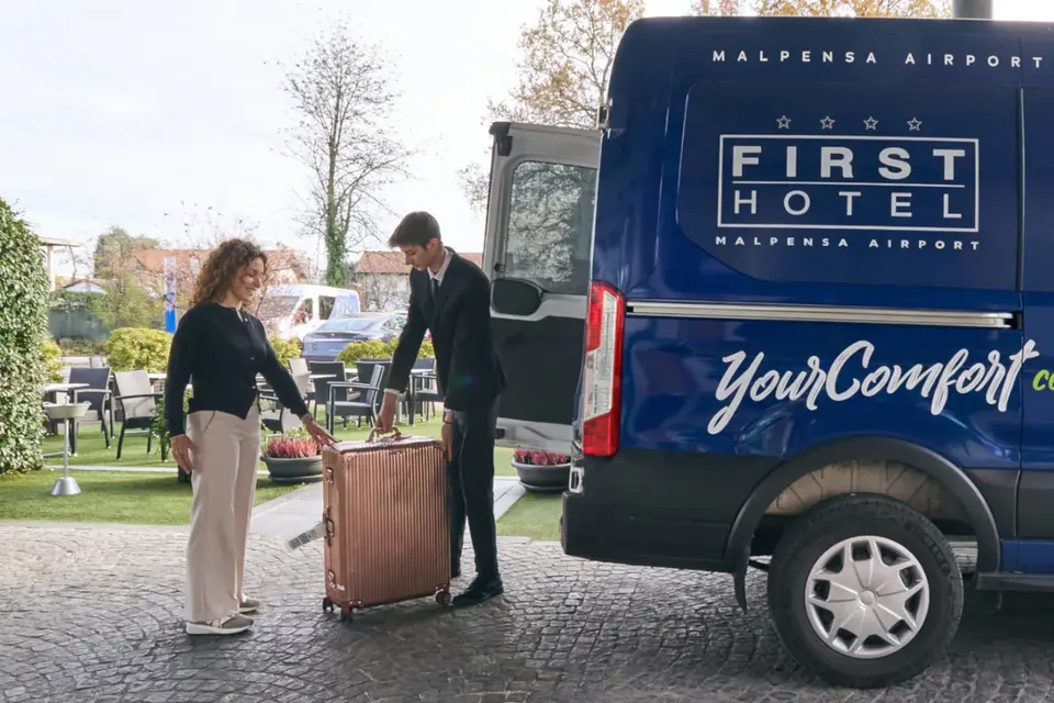 La navetta del First Hotel, blu con la scritta YourComfort comes First, davanti all’hotel: un ospite e un addetto caricano una valigia rosa